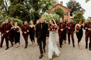 A wedding party in burgundy attire running and celebrating on a gravel path.