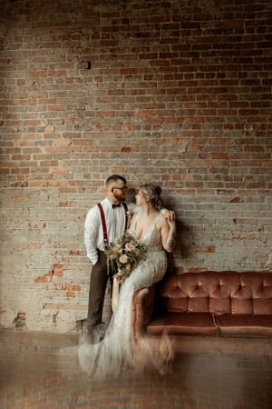 Elegant wedding couple posing by vintage sofa against exposed brick wall with natural lighting.