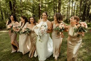 Bridesmaids in champagne dresses holding blush and white floral bouquets in a forest setting.
