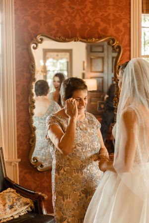 An emotional moment during wedding preparations reflected in an ornate gold mirror against red wallpaper.