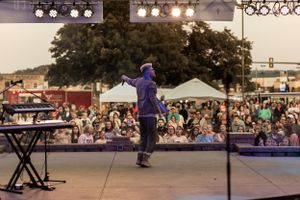 Performer in purple hoodie engaging with crowd at outdoor concert venue with stage lighting.