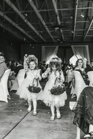 Two flower girls in white dresses carrying baskets walk down the aisle at an indoor wedding reception.