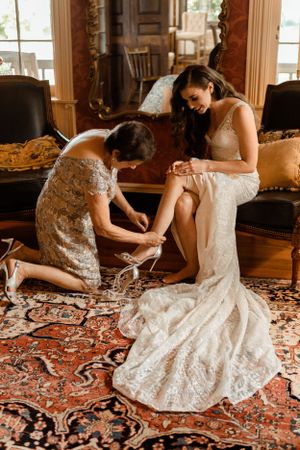 A touching wedding preparation moment showing someone helping the bride put on her shoes on an ornate Persian rug.