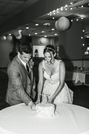 A wedding couple cuts their white wedding cake together in a reception hall decorated with string lights.