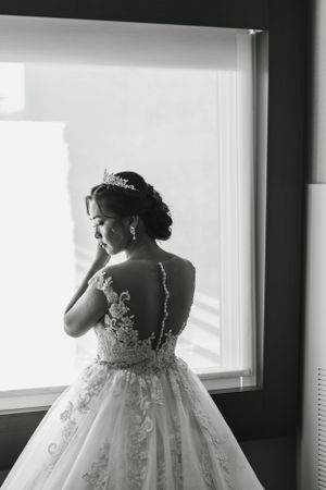 A black and white photo of a bride in a lace wedding dress looking out a window.