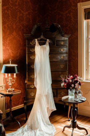 Elegant white wedding dress hanging on antique dresser in room with red damask wallpaper.