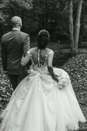 A wedding couple viewed from behind walking through a wooded area, with the bride wearing an elegant lace-back dress.