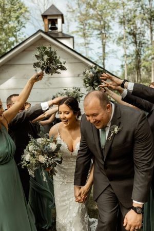 Wedding party holding bouquets overhead outside white chapel with bell tower.