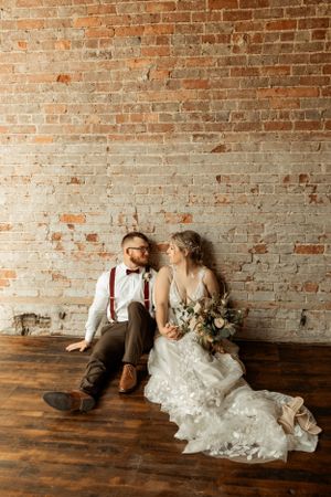 Wedding couple seated on hardwood floor against rustic exposed brick wall.