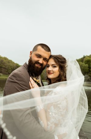 Intimate wedding portrait with couple embracing under flowing veil by water.