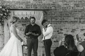 Black and white wedding ceremony photo with macrame backdrop against brick wall.