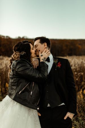 A bride wearing a leather jacket over her wedding dress shares a romantic moment with her groom in a fall field.