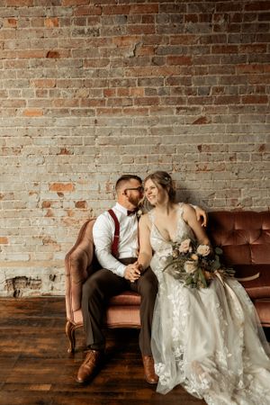 A romantic wedding photo of a couple on a vintage brown sofa against an exposed brick wall with a floral bouquet.