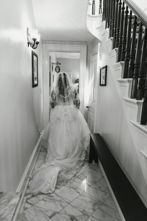 A bride in a flowing white dress walking down a narrow hallway with a staircase.