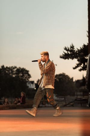 Performer in casual attire singing on outdoor stage during golden hour.
