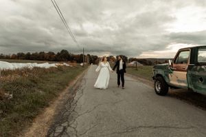 Newlyweds walk down rural road with vintage truck under dramatic cloudy sky.