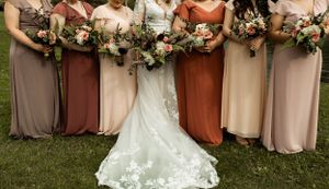 Row of bridesmaids in multicolored dresses standing with bouquets next to bride's lace train.