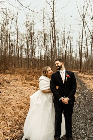 Couple in wedding attire embracing on a winter path surrounded by bare trees.