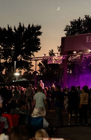 Outdoor concert venue at dusk with crescent moon visible above stage and crowd silhouettes below.