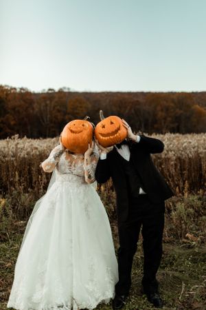 A creative wedding photo with the couple wearing jack-o-lantern pumpkins on their heads in an autumn field.