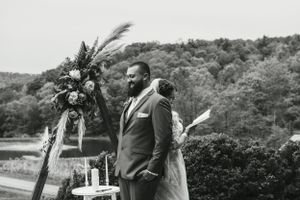 A black and white wedding photo showing a formal ceremony setup with floral arch in a scenic outdoor setting.