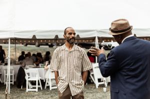 Two men having an animated conversation outdoors near white folding chairs and a tent.