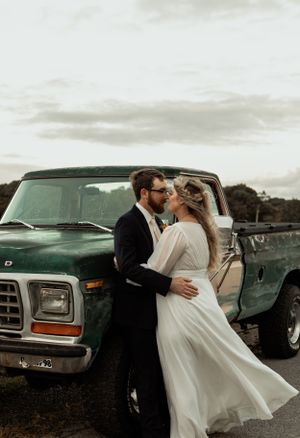 Romantic newlywed couple embraces against vintage truck during sunset photo session.