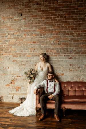 Stylish wedding portrait with a vintage sofa against rustic brick wall featuring elegant attire and bouquet.