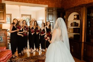 Bridesmaids in black dresses react with joy upon seeing the bride in her white gown.