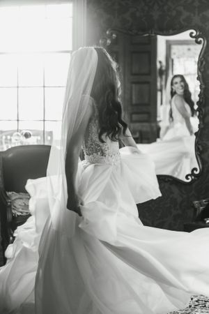 Bride in a flowing lace wedding gown twirls in front of an ornate mirror in a sunlit dressing room.