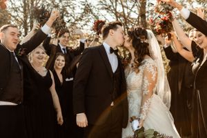 Wedding party celebrates with raised arms as newlyweds share a moment during autumn outdoor ceremony.