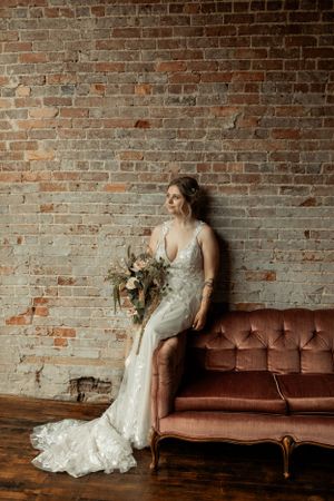 Bride in lace gown holds neutral bouquet while posing against brick wall with vintage sofa.
