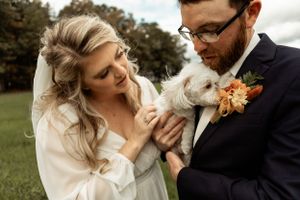 Newlyweds share a joyful moment with their small white dog wearing a floral collar at their outdoor wedding.