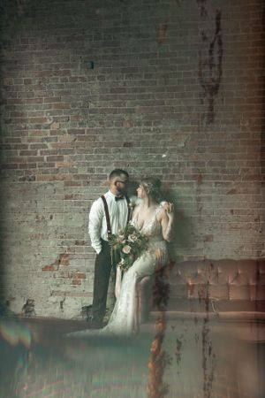Artistic wedding portrait with moody lighting against textured brick wall and vintage furniture.