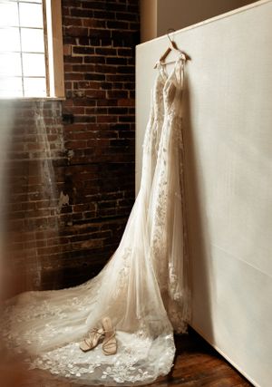 A delicate lace wedding dress hangs against a brick wall with bridal shoes beneath.