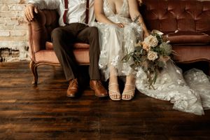 Close-up of wedding shoes and dress details with a floral bouquet on a brown vintage sofa.