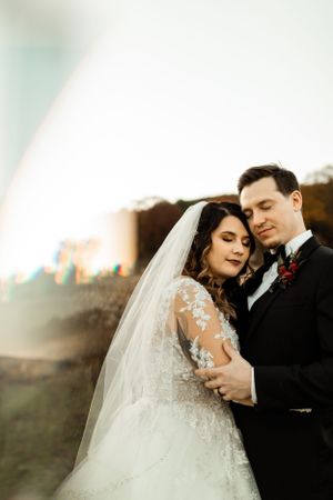 A romantic wedding portrait of newlyweds embracing at sunset with the bride in a lace-sleeved gown and flowing veil.