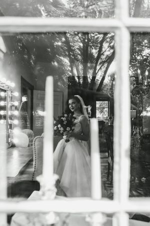 A black and white portrait of a bride through a window pane holding her bouquet.