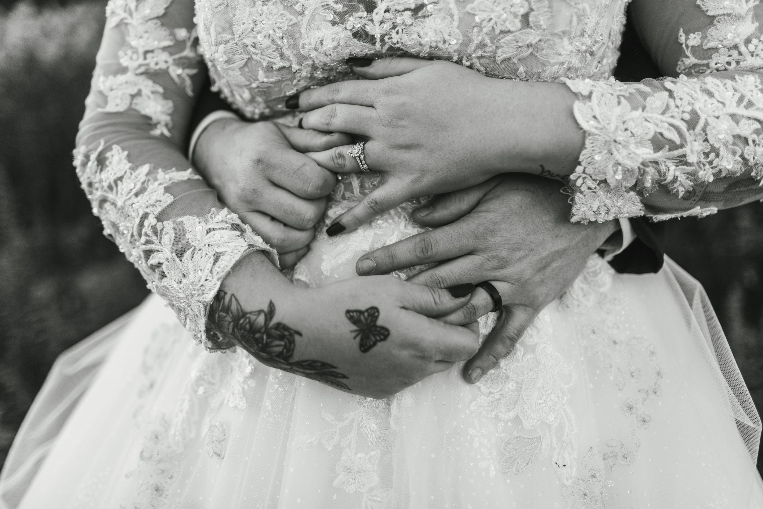 Black and white close-up of hands with wedding rings and tattoos resting on a white lace dress.