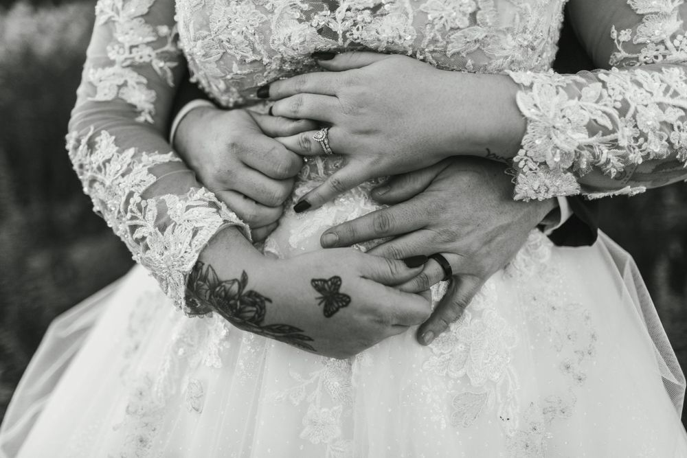 Black and white close-up of hands with wedding rings and tattoos resting on a white lace dress.