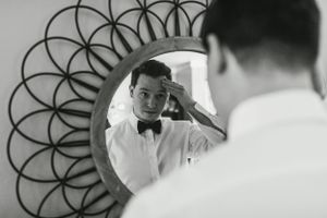 A black and white image of someone adjusting their bowtie in a decorative round mirror.