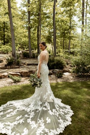 Elegant lace wedding gown with flowing train photographed in woodland garden setting.