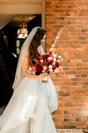 A bride in a lace wedding gown holds a stunning burgundy and pink bouquet against a brick wall.