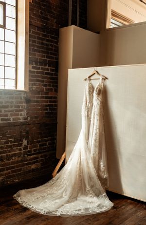 Elegant lace wedding dress hanging against exposed brick wall in natural window light.