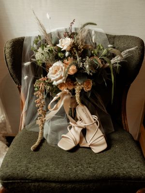 Wedding bouquet with peach roses and greenery arranged with bridal shoes on a dark green velvet chair.