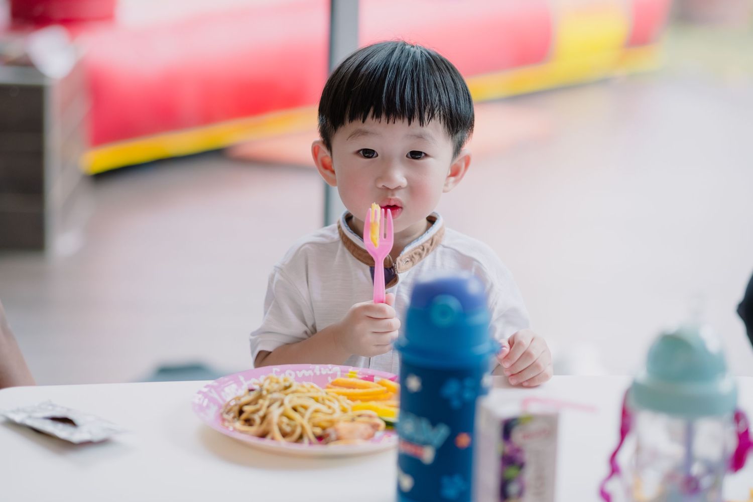 Young child enjoying noodles at a table with colorful drinks in the background.