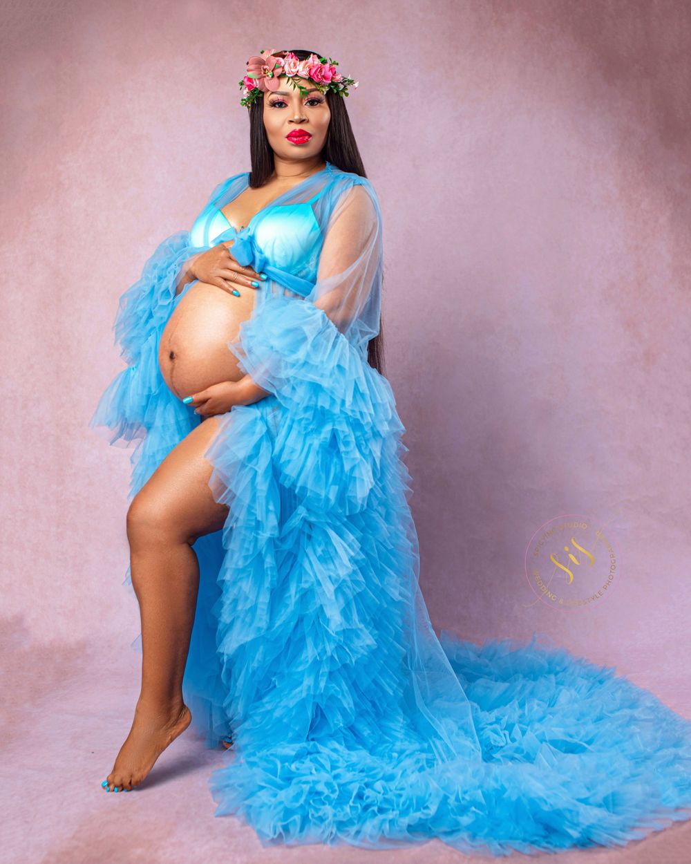 Model in ethereal bright blue tulle gown poses against pink backdrop in artistic maternity photoshoot.