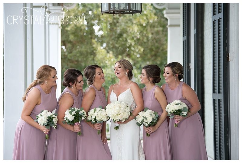 Bridesmaids in dusty rose dresses stand in line on a white columned porch.