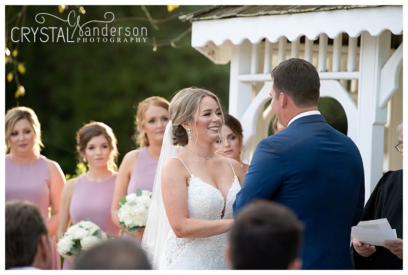 Outdoor wedding ceremony takes place under a white gazebo surrounded by natural greenery.