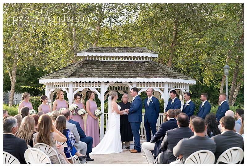 Wedding guests seated in white chairs watch ceremony under a decorative white gazebo.